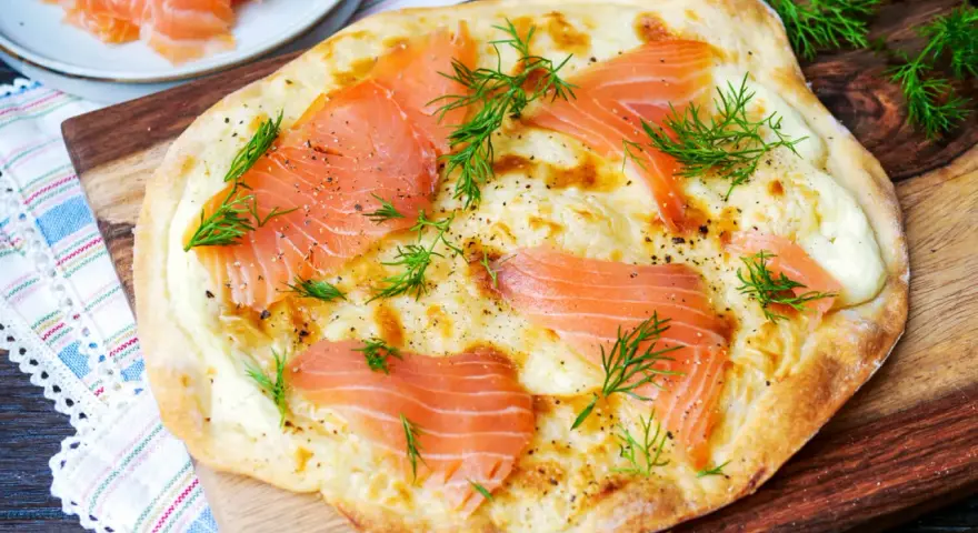 Flammkuchen mit Lachs, belegt mit zarten Lachsscheiben und frischem Dill, auf einem Holzbrett serviert.