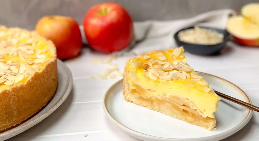 Ein saftiger Apfelkuchen mit Schmandguss, bestreut mit Mandelblättchen. Daneben liegen frische Äpfel.