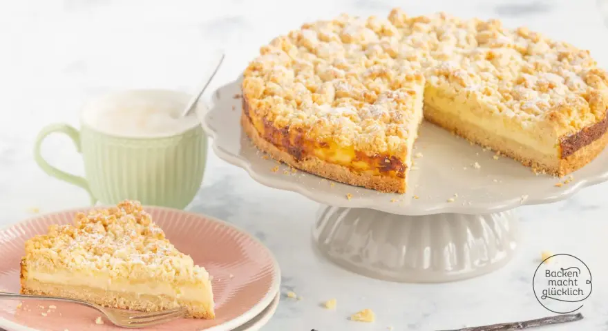 Ein saftiger Streuselkuchen mit Puddingfüllung, bestäubt mit Puderzucker. Daneben eine Tasse Cappuccino.