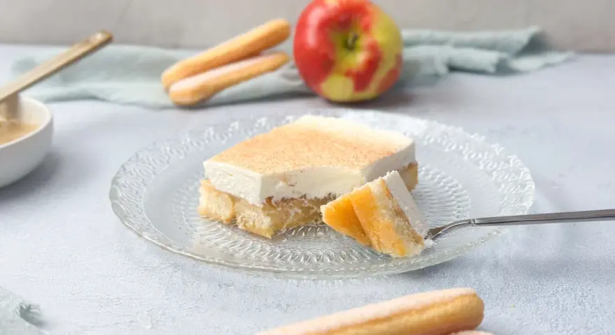 Ein Stück Apfel-Schmand-Kuchen aus der Springform, mit Löffelbiskuits und einem Apfel im Hintergrund.