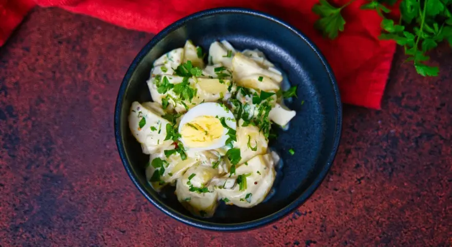 Ein cremiger Kartoffelsalat mit Mayo, garniert mit Petersilie und einem halben Ei, serviert in einer dunklen Schüssel.
