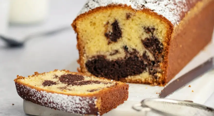 Ein saftiger marmorkuchen mit Puderzucker bestäubt, daneben ein Stück Kuchen und ein Sieb. Perfekt mit einem Klecks Sahne.
