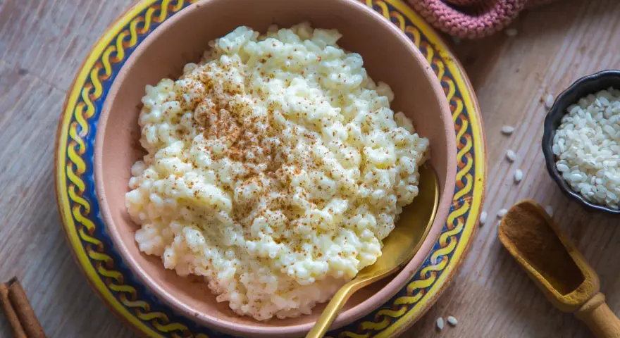 Cremiger Milchreis, bestreut mit Zimt, wartet in einer Schale auf sein Milchreis Rezept. Daneben liegen Reiskörner und Zimtstangen.
