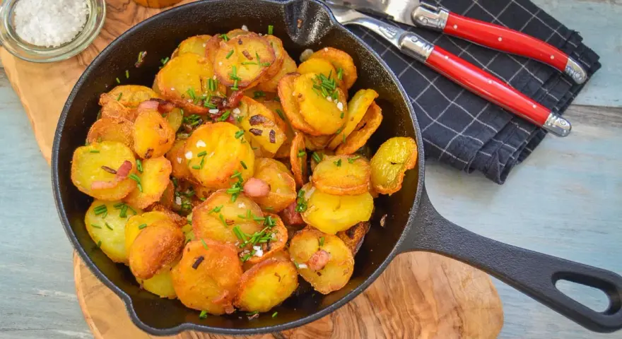 Bratkartoffeln mit Zwiebeln und Speck in einer Gusseisenpfanne, garniert mit Schnittlauch. Dazu grobes Salz und ein Messer-Gabel-Set.