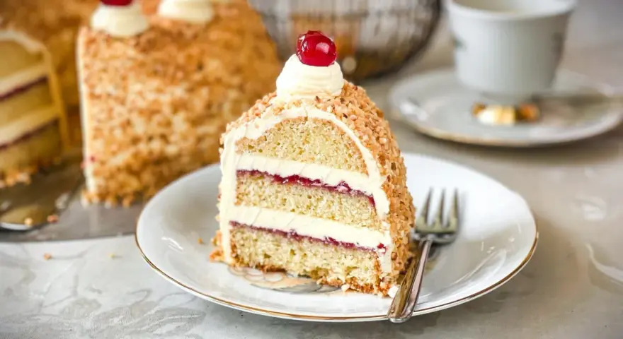 Ein Stück Frankfurter Kranz, das Rezept eines Konditors, mit Buttercreme und Kirschen verziert, umgeben von gehackten Nüssen.