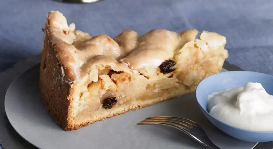 Ein Stück gedeckter Apfelkuchen mit Rosinen und Zuckerguss, serviert mit einem Klecks Sahne.