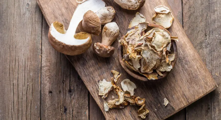 Frische und getrocknete Steinpilze liegen auf einem Holzbrett. Die getrockneten Pilze sind in einer Schale und daneben verteilt, bereit zum Kochen.