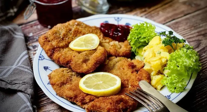Knuspriges Wiener Schnitzel mit Zitrone, Preiselbeeren und Kartoffelsalat auf einem Teller.
