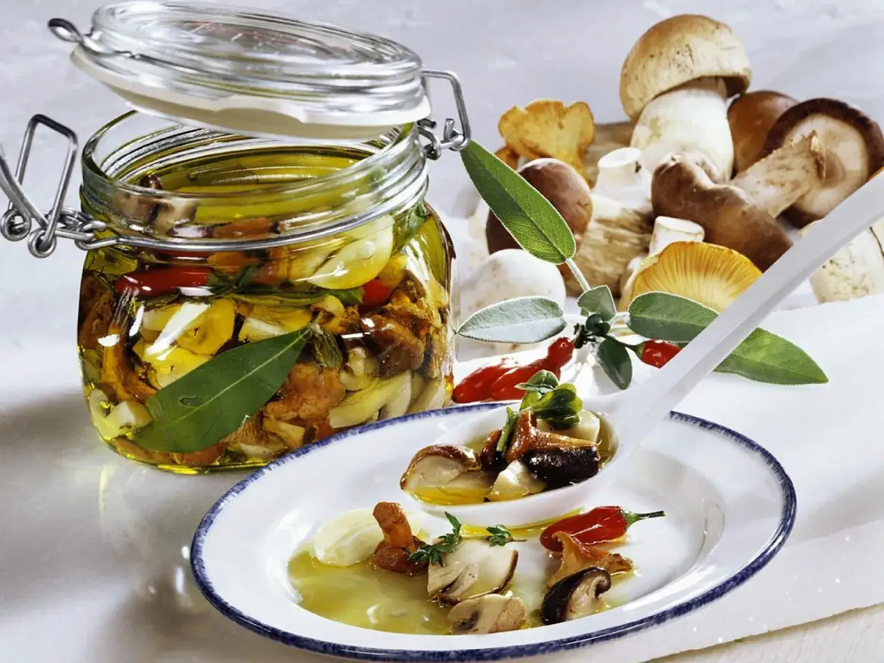 Frische Pilze, Knoblauch und Kräuter in Öl eingelegt. Ein Glas mit eingelegten Pilzen und eine Löffelportion auf einem Teller zeigen die Zubereitung von pilze einlegen.