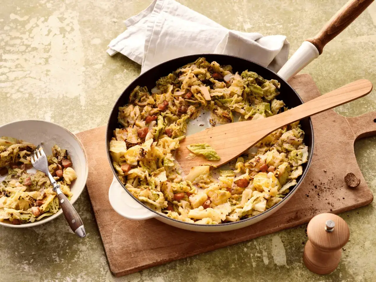 Herzhafte Wirzing-Rezepte: Gebratener Wirsing mit Speck in einer Pfanne, daneben eine Schüssel mit einer Gabel.