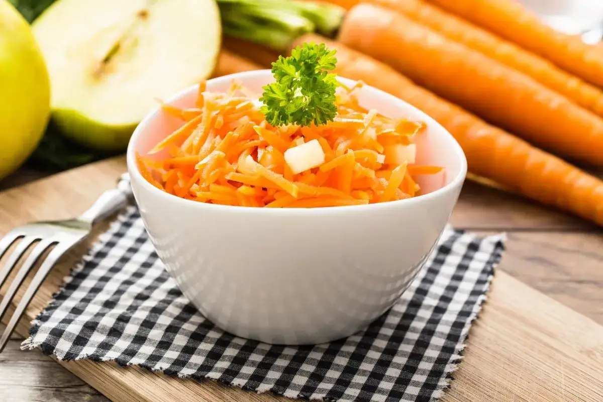 Frischer Karotten-Apfel-Salat, garniert mit Petersilie, neben ganzen Karotten und Apfelh&auml;lften.