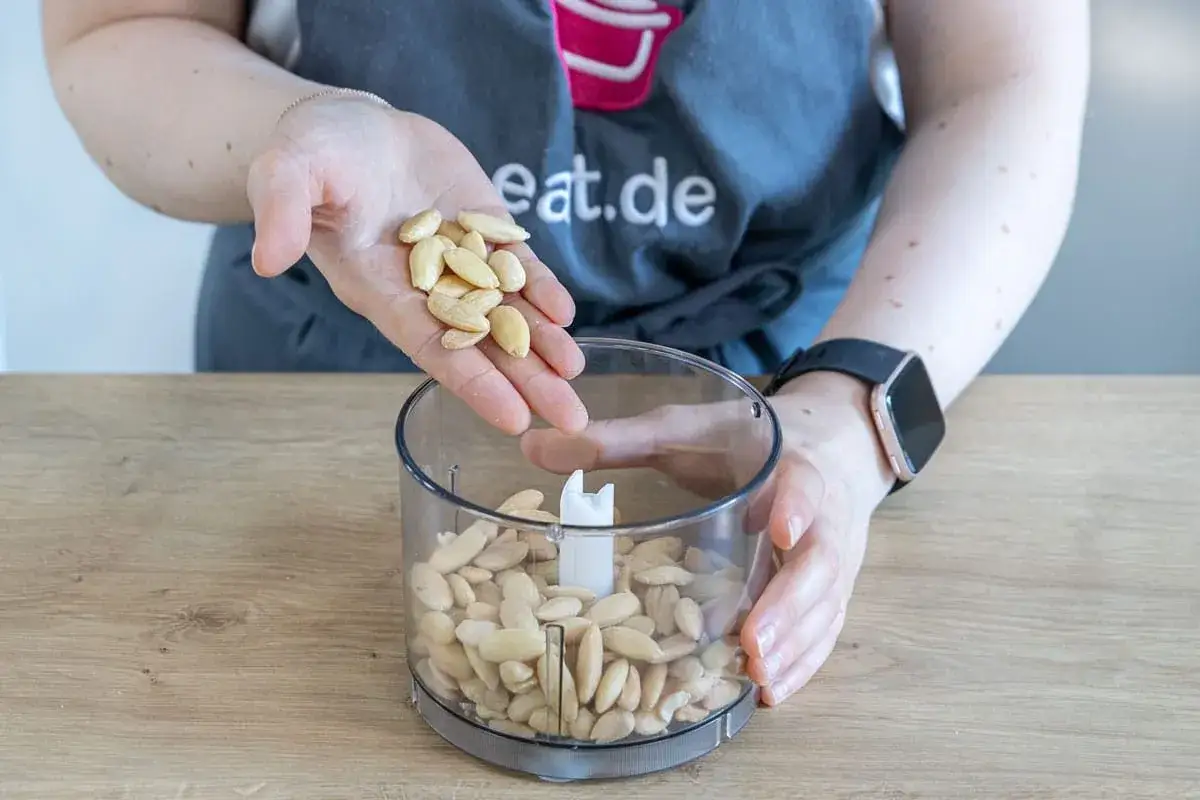 Hände geben blanchierte Mandeln in einen Mixer, um Marzipan selbst zu machen, ganz ohne Rosenwasser.