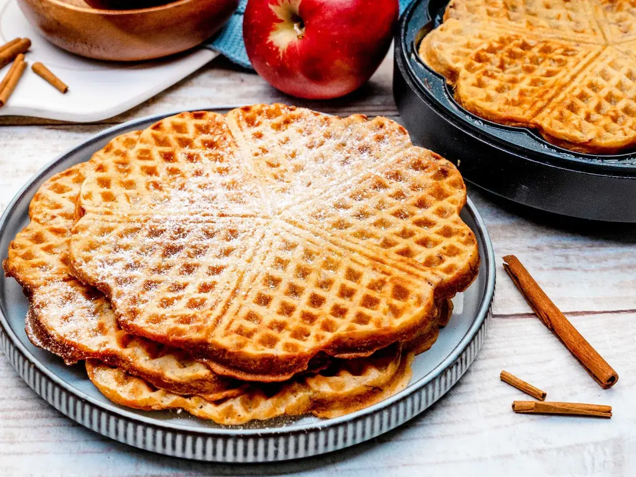 Herzförmige Waffeln mit Puderzucker bestäubt, daneben ein roter Apfel und Zimtstangen. Perfekt für einen gemütlichen Nachmittag mit waffeln mit apfel.