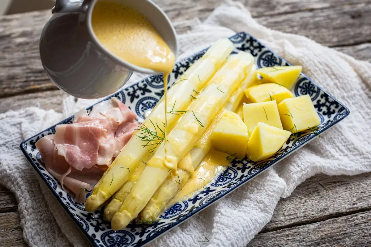 Spargel mit Schinken und Kartoffeln, übergossen mit Sauce Hollandaise. Ein klassisches Frühlingsgericht, das auf einem rustikalen Holztisch serviert wird.