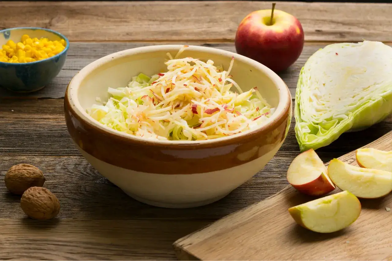 Schüssel mit geraspeltem Spitzkohl und Apfel, bereit für ein einfaches spitzkohlsalat rezept. Daneben liegen Walnüsse, Mais und Apfelstücke.