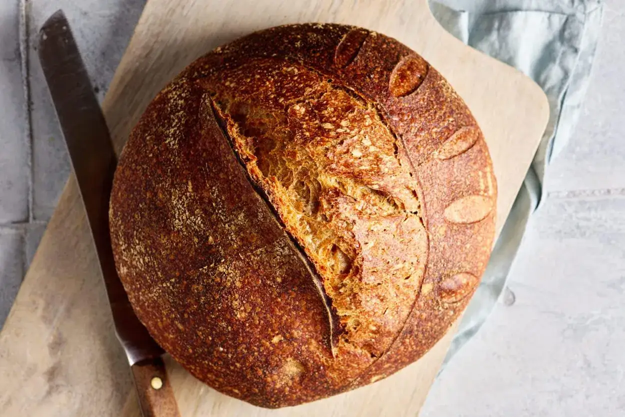 Ein frisch gebackenes Dinkelbrot mit knuspriger Kruste liegt auf einem Holzbrett. Daneben ein Messer und ein hellblaues Tuch.