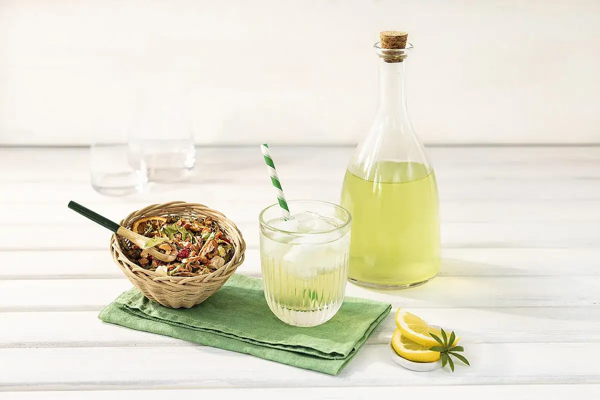 Erfrischende Maibowle: Ein Glas mit Eis, eine Flasche grüner Flüssigkeit, Kräuter in einem Körbchen und Zitronenscheiben – perfekt für ein Maibowle Rezept.