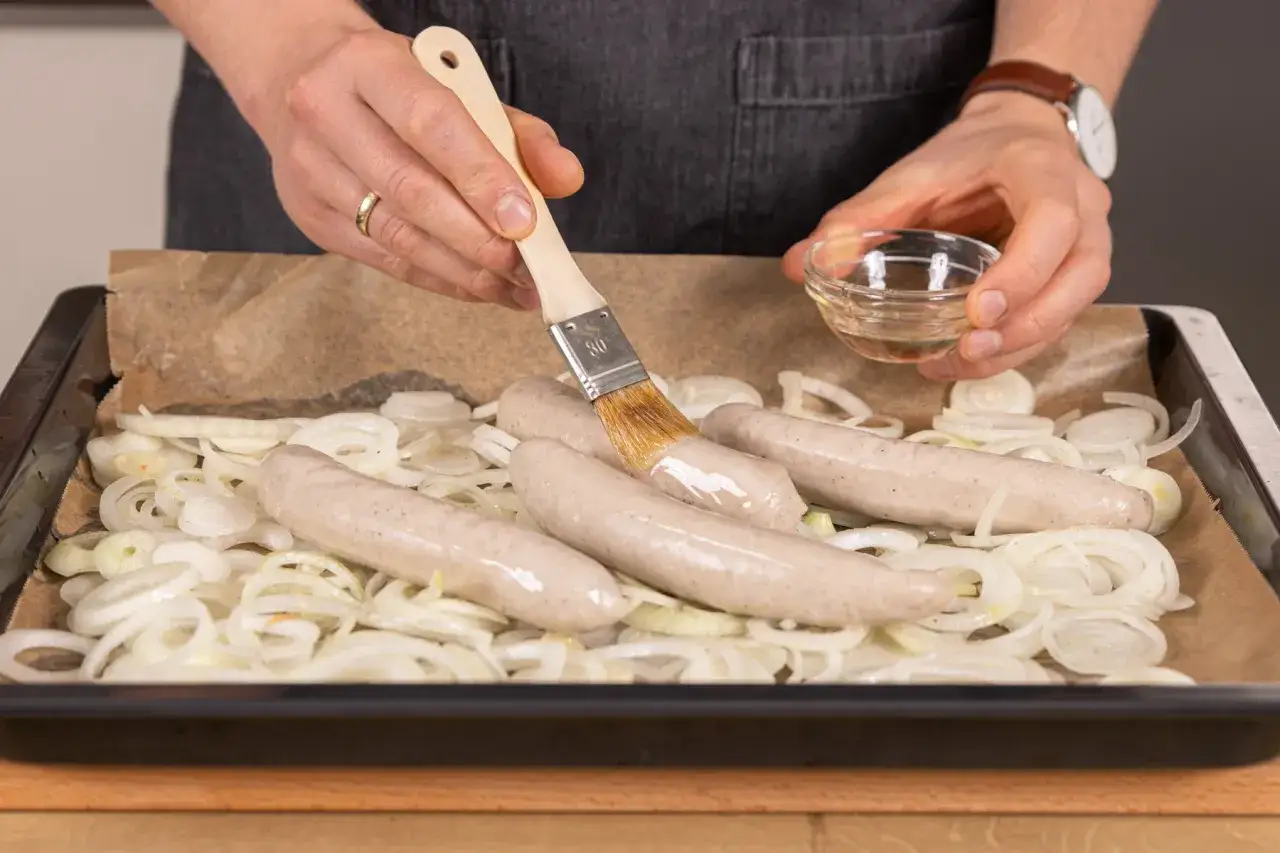 Bratwurst im Backofen: Zwiebelringe auf Backpapier, darauf vier rohe Bratwürste. Mit einem Pinsel wird eine Flüssigkeit auf die Würste aufgetragen.