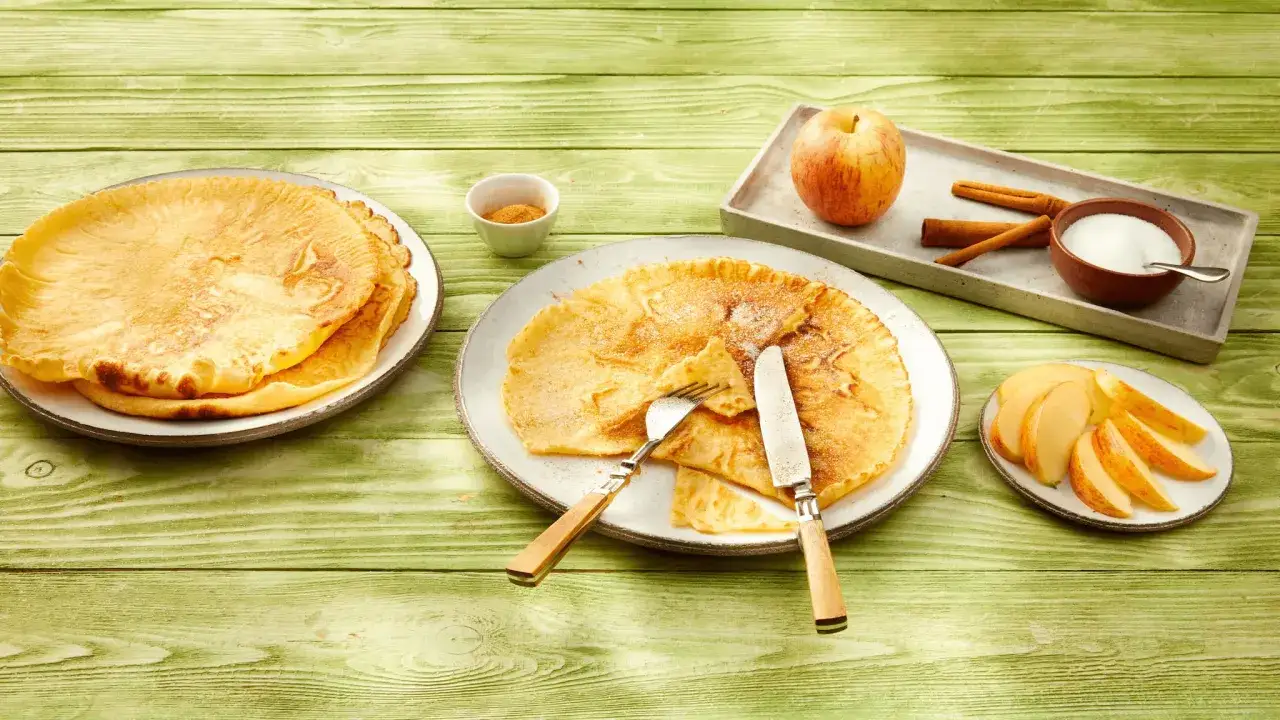 Ein leckeres Pfannkuchen Rezept: goldbraune Pfannkuchen auf Tellern, dazu Apfelschnitze, Zimt und Zucker. Perfekt für ein süßes Frühstück!