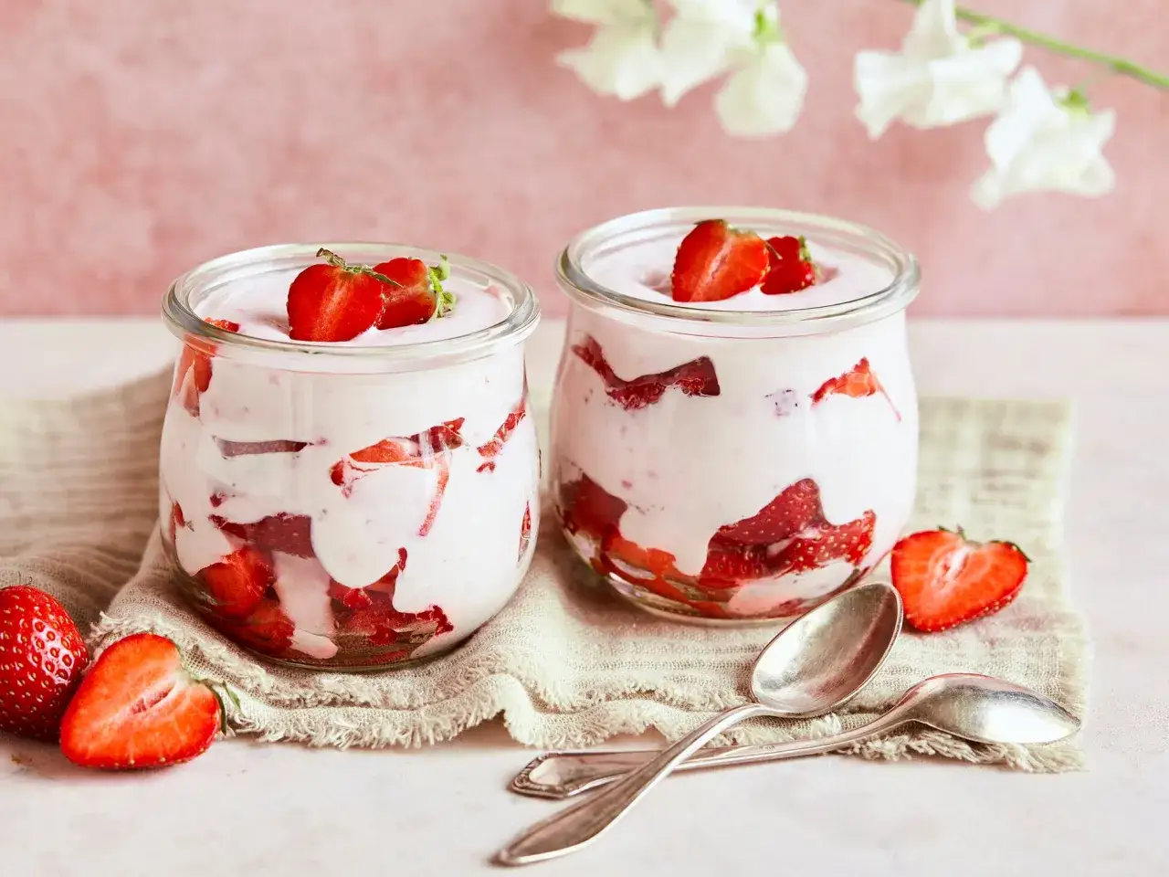 Zwei Gläser mit cremigem Erdbeer Quark Dessert, garniert mit frischen Erdbeeren. Löffel liegen daneben.