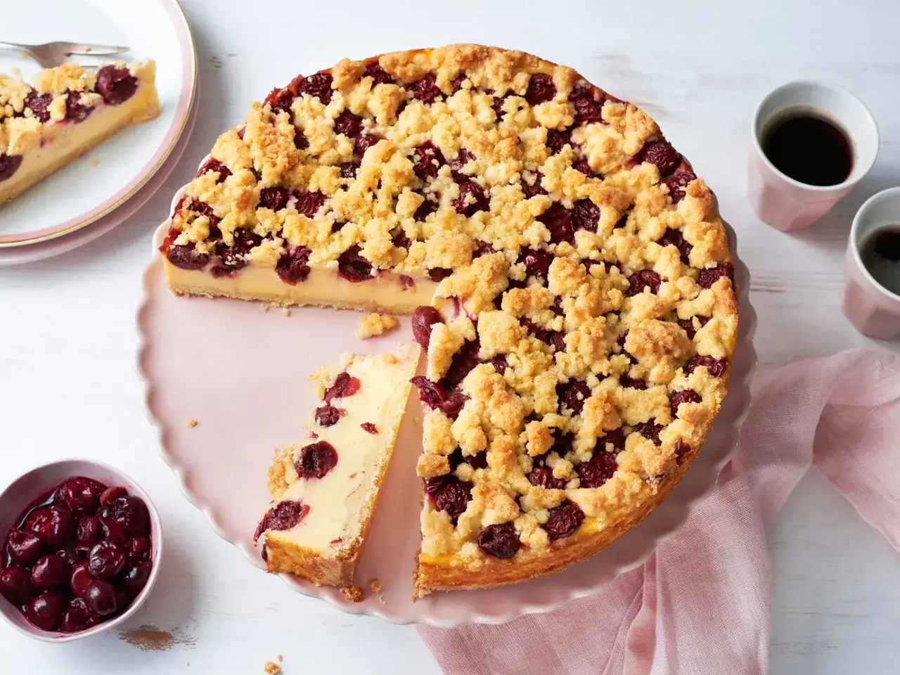 Ein saftiger Pudding Kirsch Kuchen mit Streuseln, daneben eine Schale Kirschen und zwei Tassen Kaffee.
