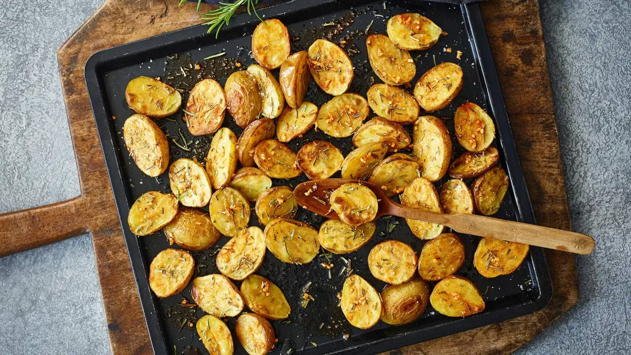 Goldbraune Drillinge im Ofen, gew&uuml;rzt mit Rosmarin und Knoblauch, liegen auf einem Backblech. Ein Holzl&ouml;ffel hebt eine Kartoffel an.