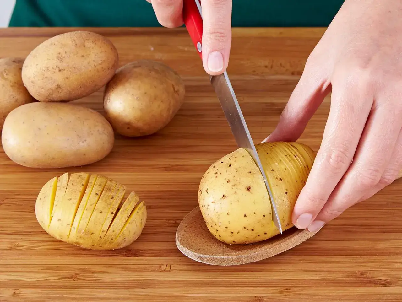 Hasselback Kartoffeln werden vorbereitet: Eine Kartoffel wird auf einem Holzl&ouml;ffel liegend mit einem Messer eingeschnitten.
