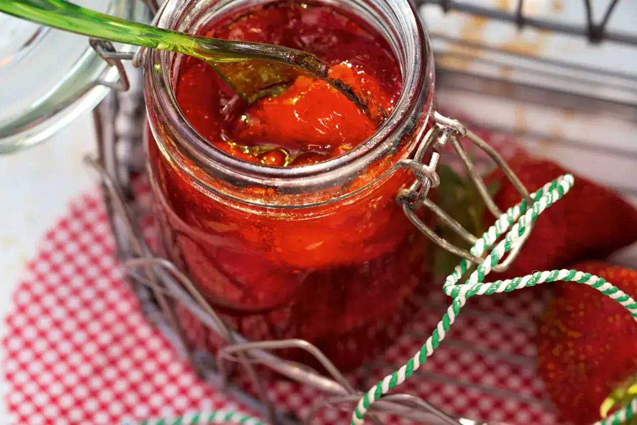 Ein Glas mit leuchtend roter Erdbeermarmelade, die mit einem grünen Löffel probiert wird. Frische Erdbeeren liegen daneben. Diese Erdbeermarmelade mit Pfiff ist ein Genuss!