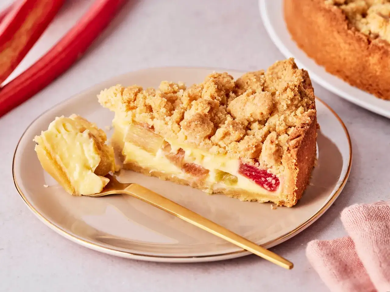 Ein Stück von Omas Rhabarberkuchen mit Pudding und Streuseln auf einem Teller, daneben ein goldener Löffel und frische Rhabarberstangen.