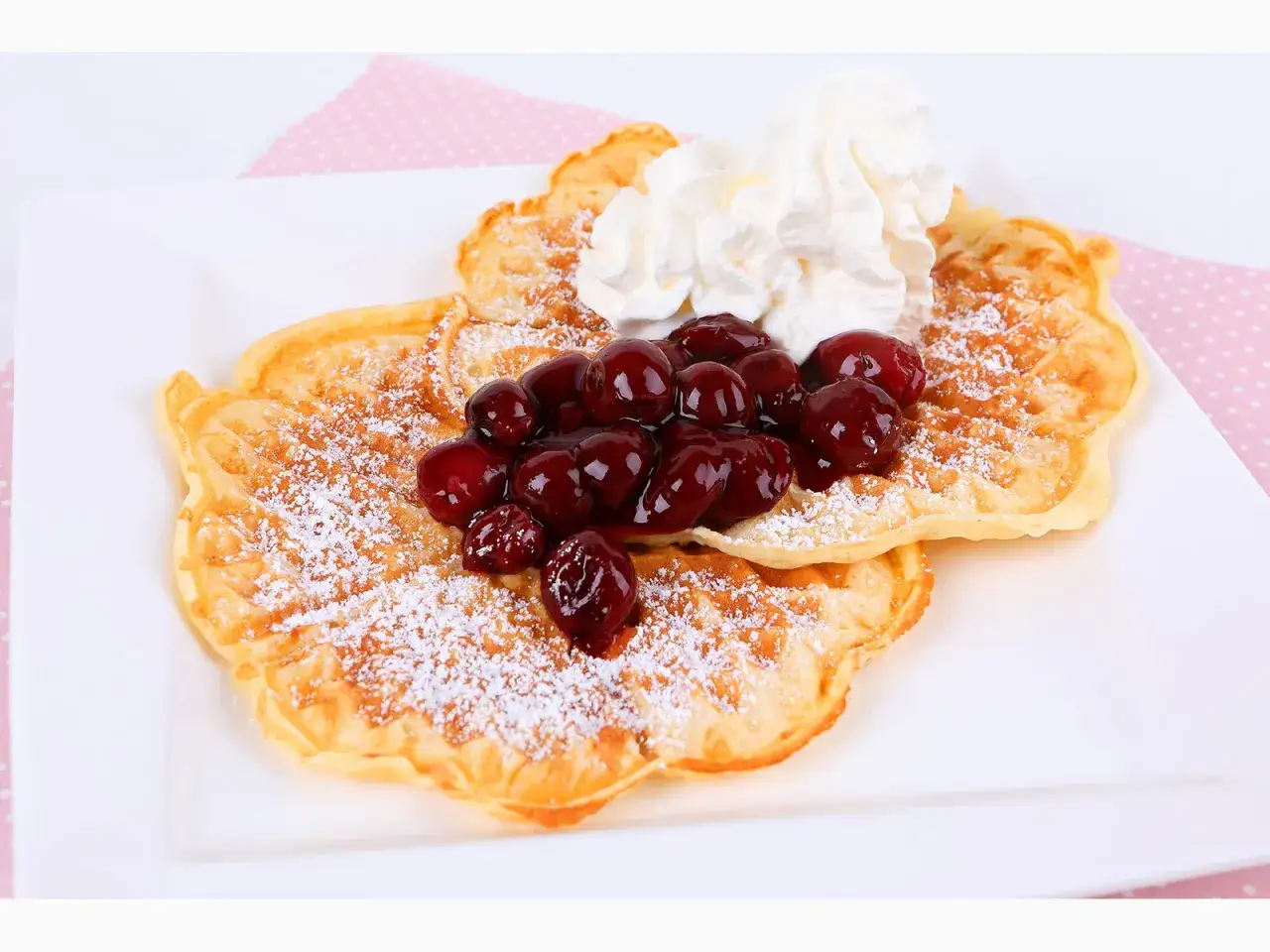 Zwei goldbraune Waffeln, bestäubt mit Puderzucker, belegt mit Kirschen und Sahne. Ein köstliches Waffeln Rezept für Genießer.