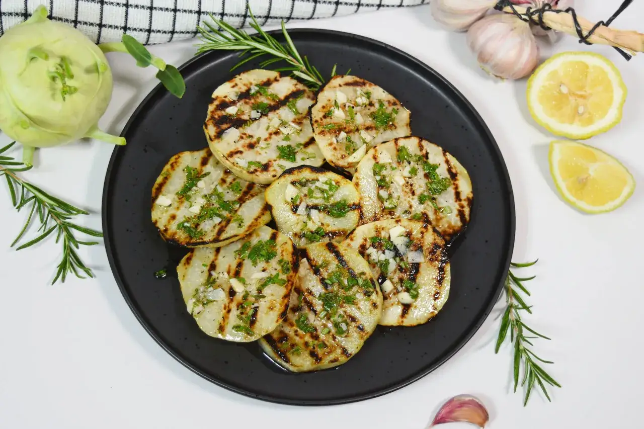 Gegrillte Kohlrabi-Scheiben mit Kräutern und Knoblauch auf schwarzer Platte, garniert mit Rosmarin und Zitrone.
