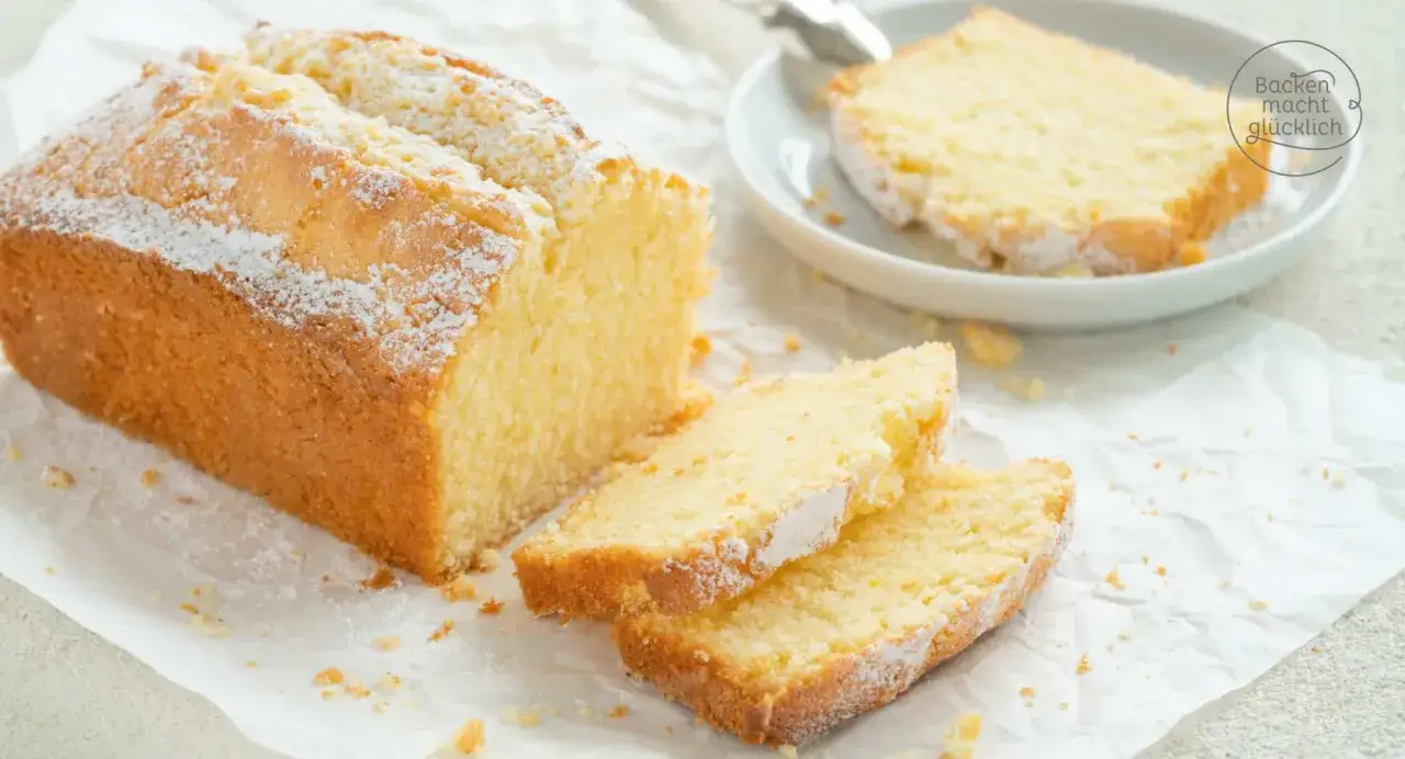 Ein goldbrauner Kastenkuchen, mit Puderzucker bestäubt, liegt auf Backpapier. Daneben liegen zwei Scheiben, die Lust auf weitere Kastenkuchen Rezepte machen.