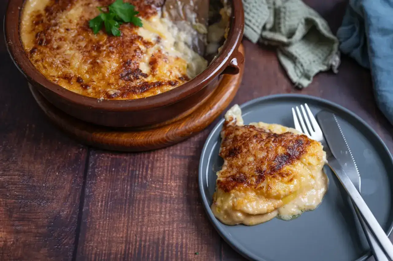 Ein goldbraun überbackenes Kartoffelgratin-Rezept, serviert in einer Terrakotta-Schüssel und auf einem Teller mit Messer und Gabel.