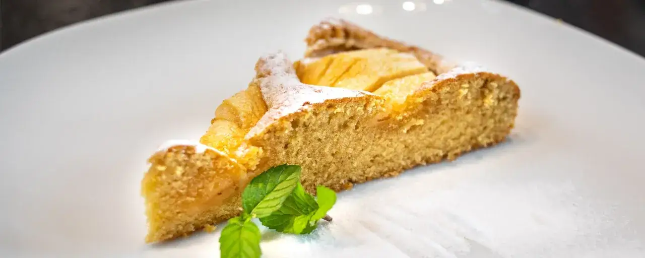 Ein Stück apfelkuchen sehr fein, mit Puderzucker bestäubt und einem Minzblatt garniert, auf einem weißen Teller.