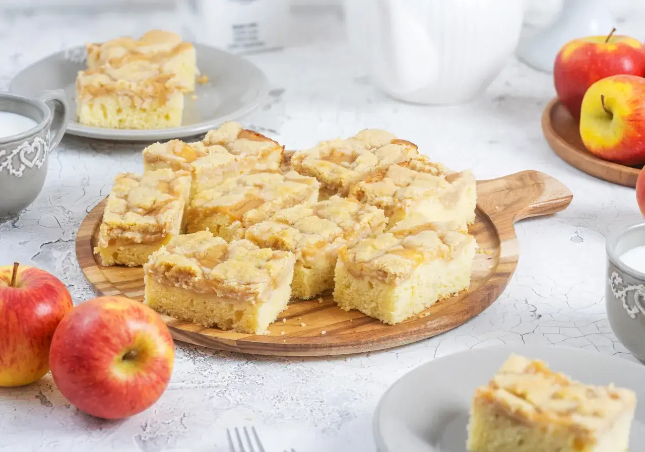 Saftiger Kuchen mit Apfelmus und Streuseln, serviert auf einem Holzbrett. Daneben Äpfel und eine Tasse Milch.
