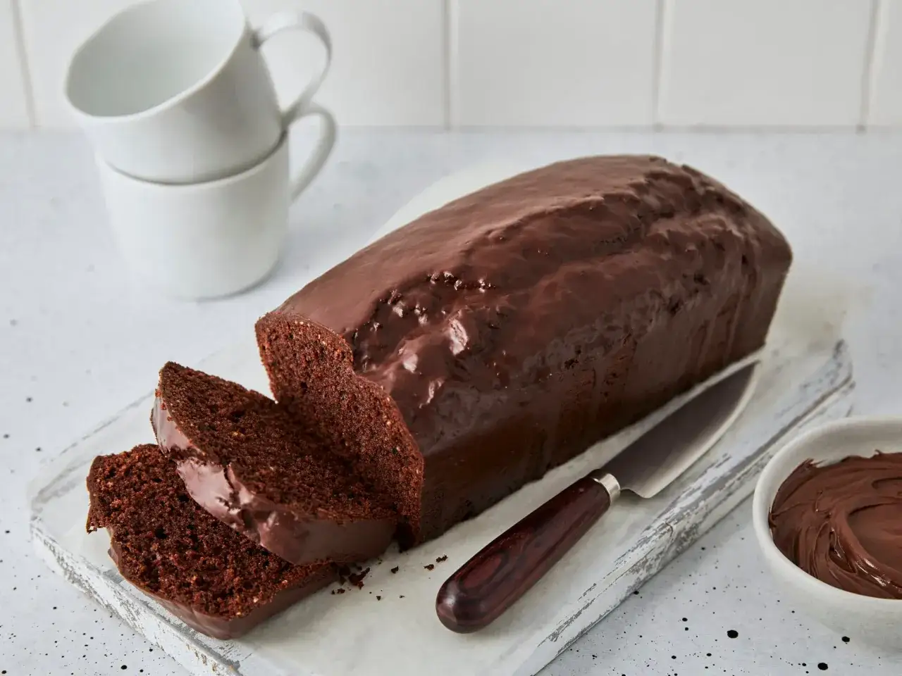 Saftiger Schokoladenkuchen mit glänzender Glasur, angeschnitten, daneben eine Schale Nutella. Perfekt für den Kaffeetisch.