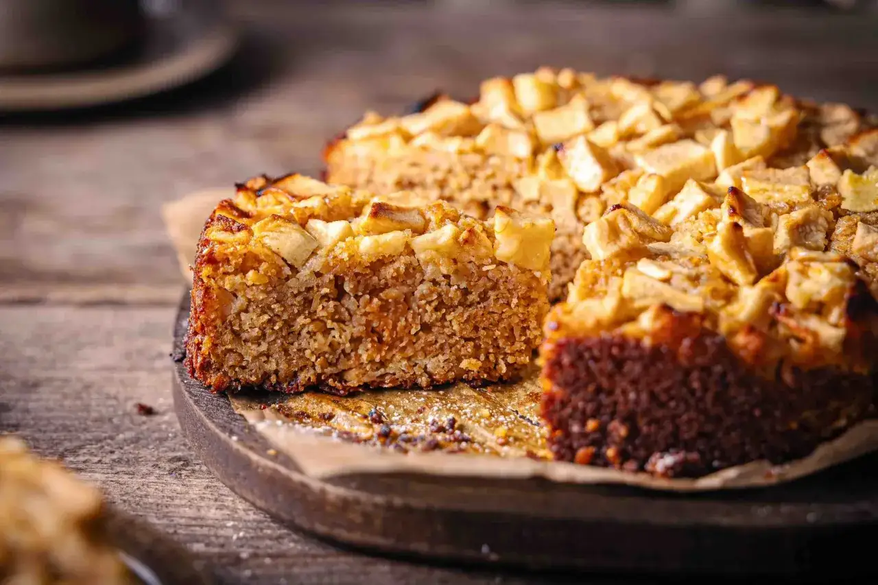 Ein saftiger Apfel-Haferflocken-Kuchen mit goldbrauner Kruste und Apfelstücken obendrauf, auf einem Holzbrett serviert.