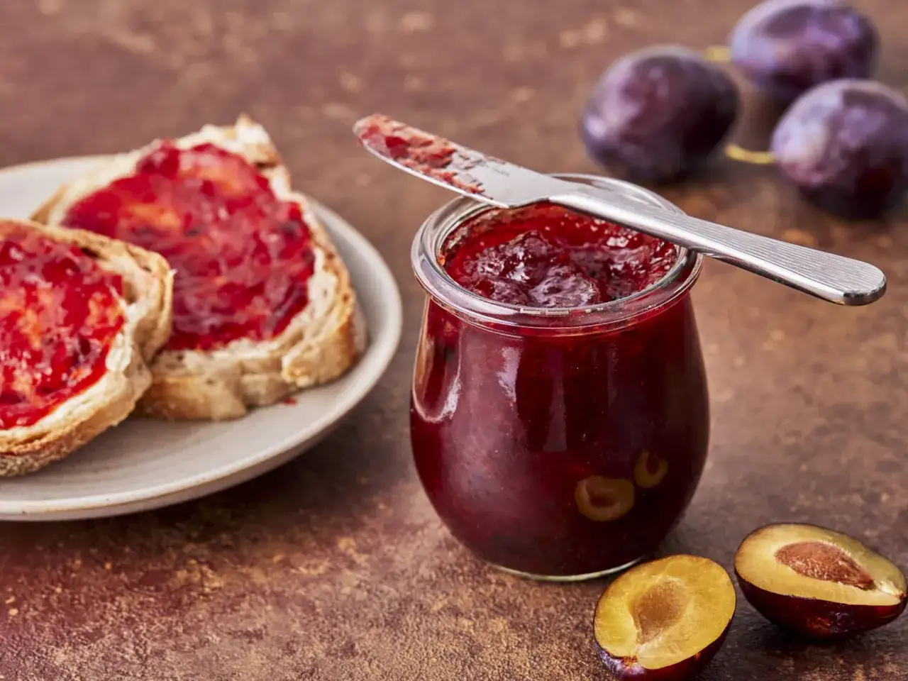 Glas mit Pflaumenmarmelade, daneben Brot mit Marmelade und frische Pflaumen. Perfekt für ein pflaumenmarmelade rezept.