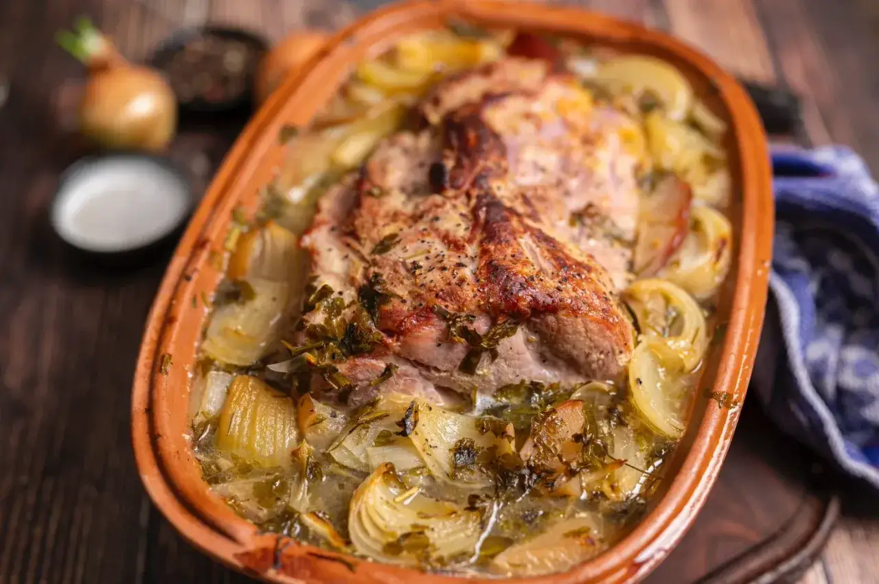 Saftiger Schweinenacken im Bräter, umgeben von Zwiebeln und Kräutern. Ein köstliches Schweinenacken Rezept, das Appetit macht.