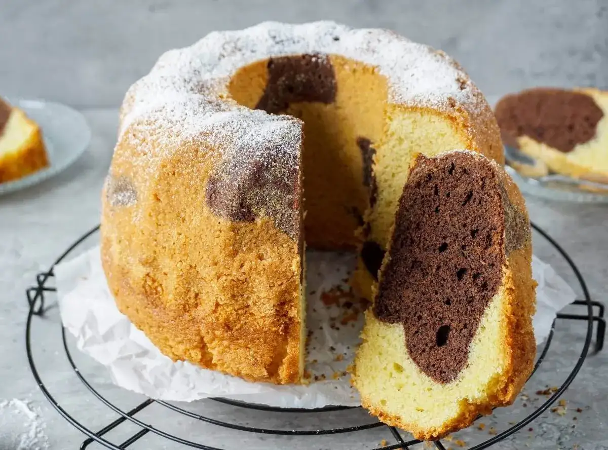 Ein saftiger Marmorkuchen, fluffig und mit Puderzucker bestäubt, wartet darauf, genossen zu werden. Ein Stück ist bereits angeschnitten.