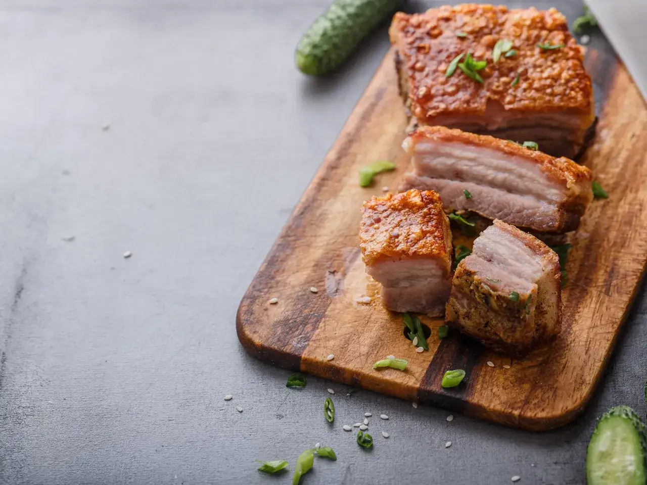 Knuspriger Schweinebauch, perfekt für ein leckeres Schweinebauch Rezept. Serviert auf einem Holzbrett mit Gurken und Frühlingszwiebeln.