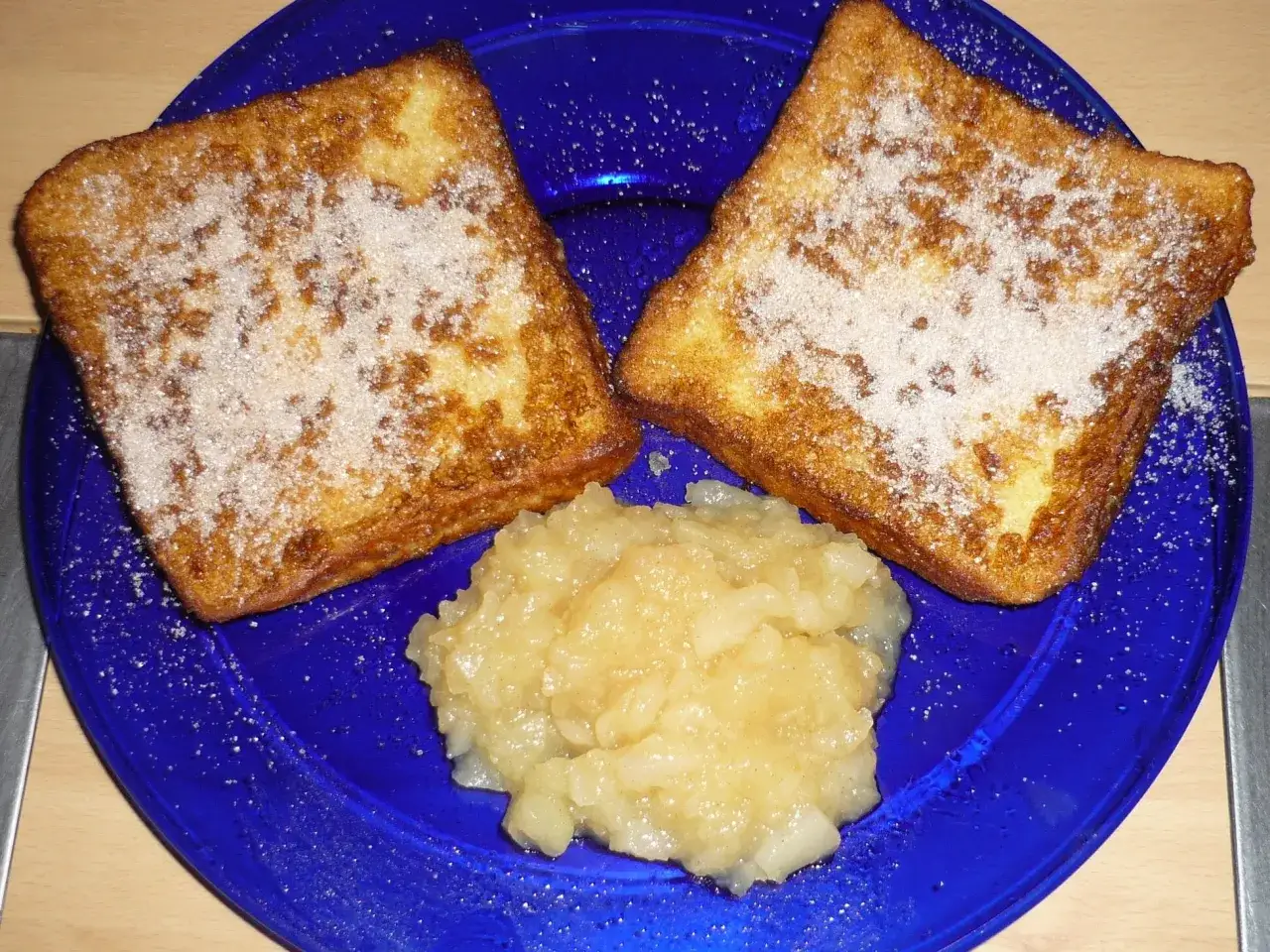 Zwei goldbraune Scheiben Toast, bestreut mit Zucker, liegen neben einem Klecks Apfelmus auf einem blauen Teller. Ein wahrhaft
