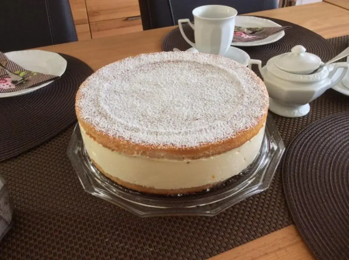 Eine köstliche Quark Sahne Torte, bestäubt mit Puderzucker, steht auf einem Tisch. Daneben eine Tasse und ein Zuckertopf.