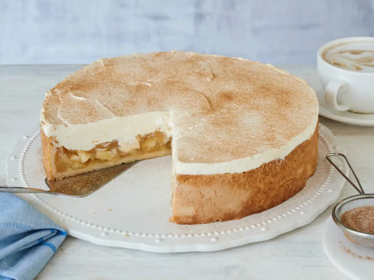 Ein saftiger Apfel-Schmand-Kuchen in Springform, bestäubt mit Zimt. Daneben eine Tasse Cappuccino.