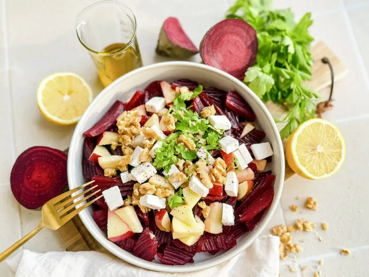 Ein bunter Rotkohlsalat mit Äpfeln, Feta und Walnüssen, garniert mit Petersilie. Daneben liegen frische Rote Bete, Zitronenspalten und ein Glas Öl.