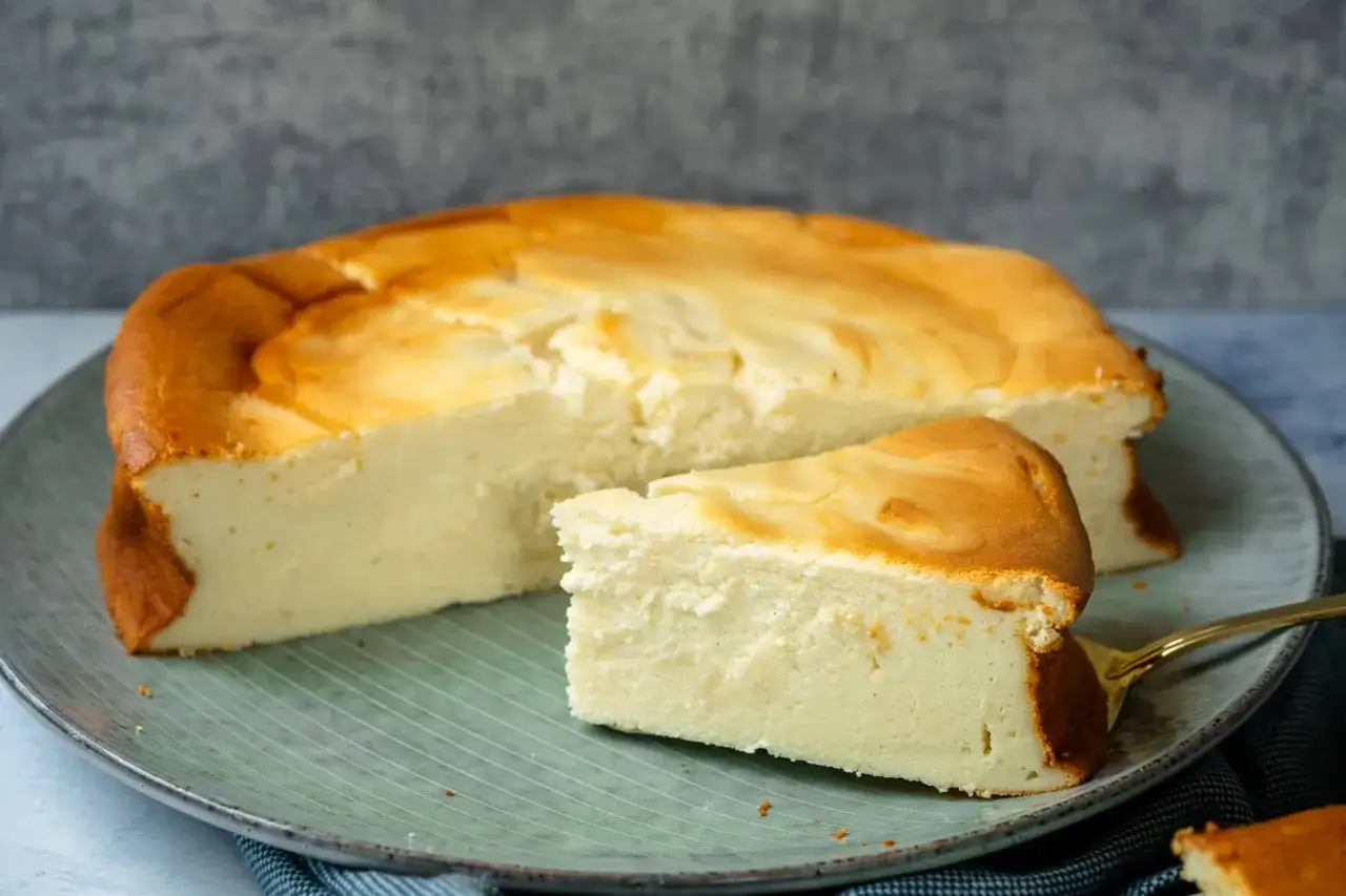Ein saftiger Quarkkuchen ohne Boden, goldbraun gebacken, liegt auf einem Teller. Ein Stück ist bereits angeschnitten und zeigt die cremige Konsistenz.