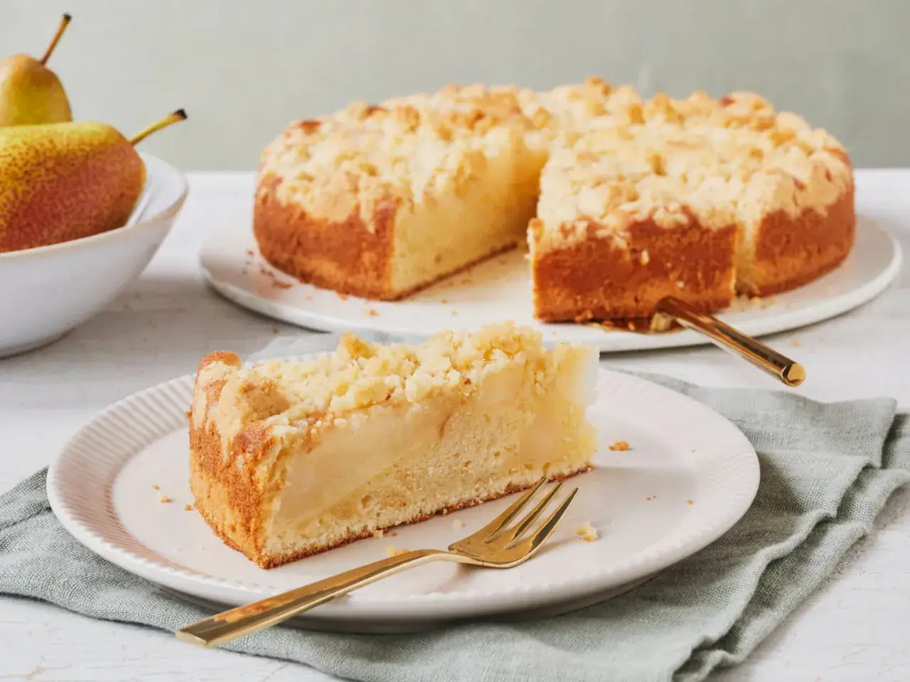 Ein saftiger Apfel-Birnen-Kuchen mit Streuseln, ein Stück davon auf einem Teller mit goldenem Besteck.