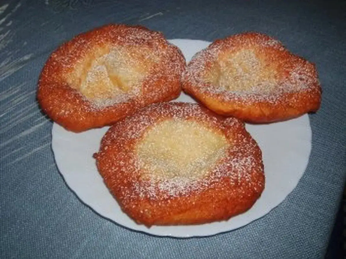 Drei goldbraune, ausgezogene Krapfen, bestäubt mit Puderzucker, liegen auf einem weißen Teller.