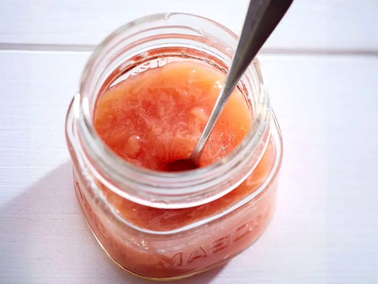Ein Glas Rhabarber-Bananen-Marmelade mit einem Löffel darin. Die Marmelade hat eine schöne rosa Farbe und eine leicht stückige Konsistenz.