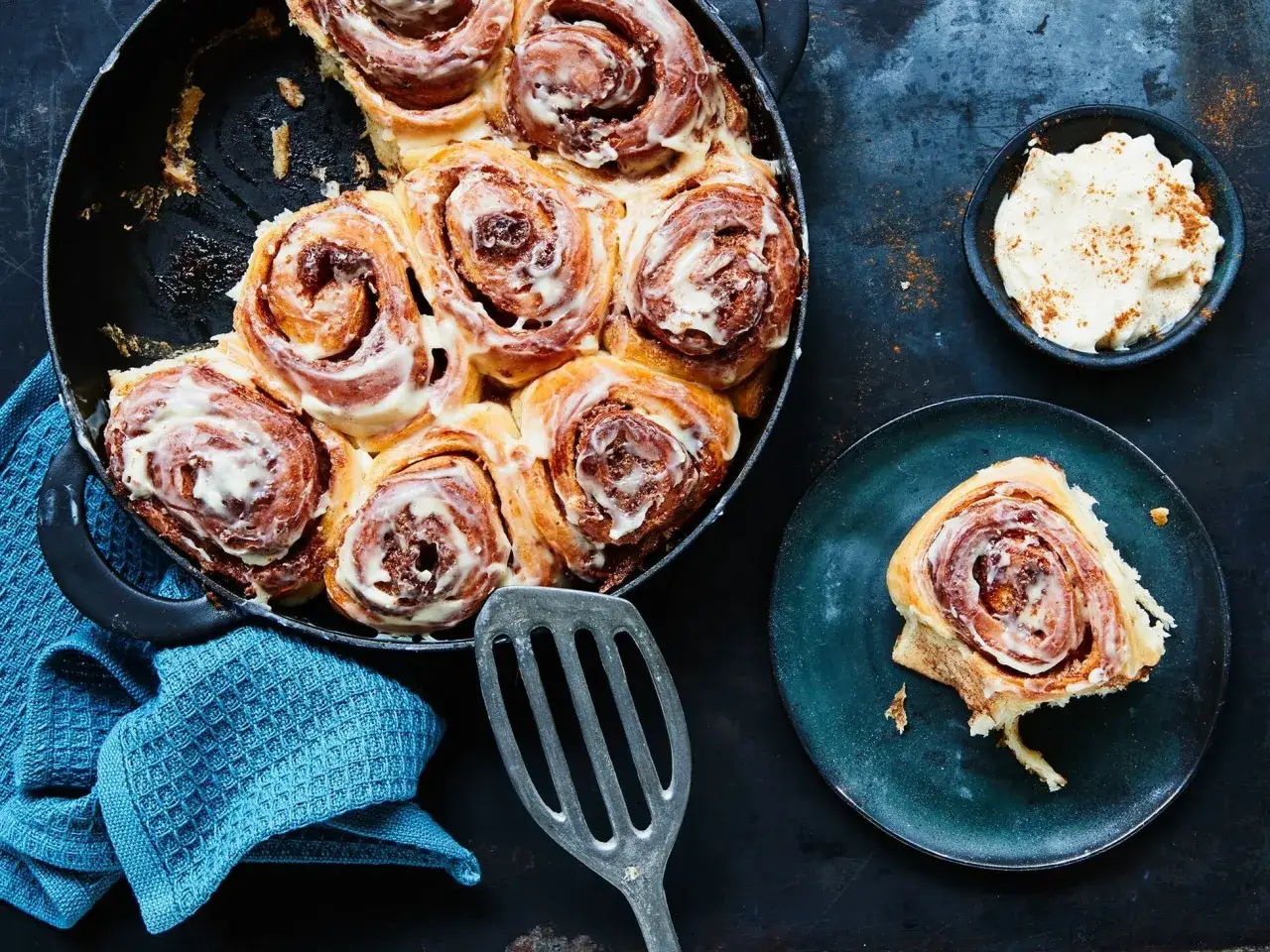 Frisch gebackene Zimtschnecken im Gusseisenpfännchen, ein köstliches Zimtschnecken Rezept, mit Zuckerguss und einer Schüssel Frischkäse-Frosting daneben.